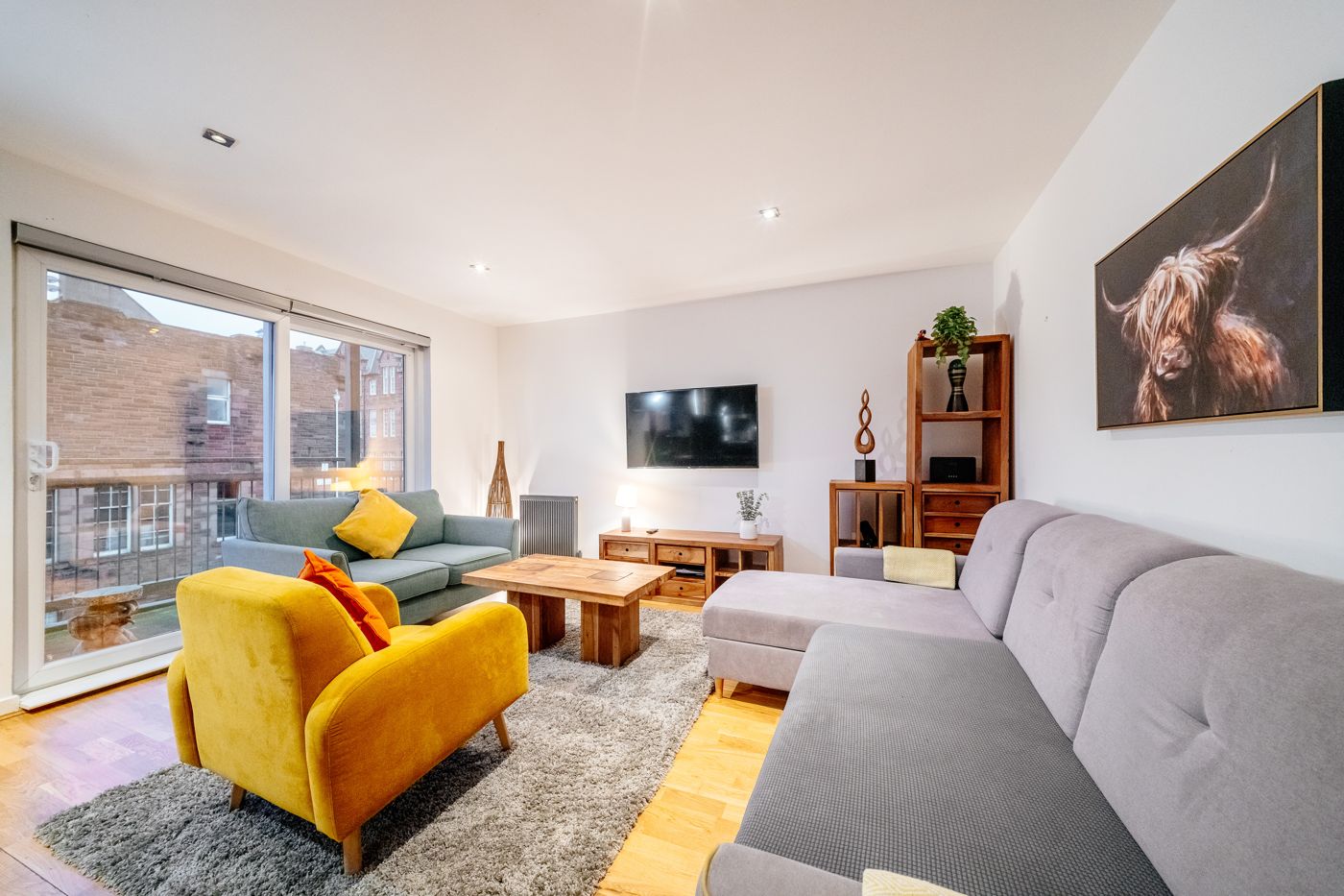 Bright living room with a yellow armchair, corner sofa and a Highland cow painting, in a home we look after near Easter Road